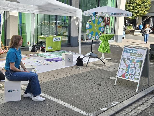 EIne VCD-Ehrenamtliche sitzt auf einem Papphocker vor dem fertig aufgebauten Infostand.