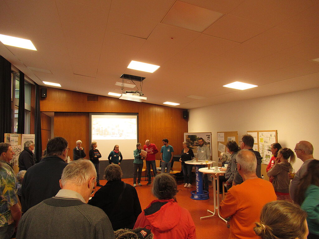 Ein Foto vom Neujahrsempfang vom VCD Freiburg. Zu sehen sind Redebeiträge von den Aktiven der Ortsgruppe Freiburg und Stellwände mit Informationen zu laufenden Projekten. Die vielen Anwesenden hören den Redebeiträgen zu.