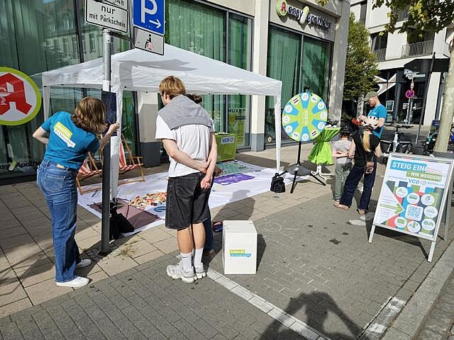 Eine VCD-Aktive und zwei Interessierte begucken vorgebeugt und nah dran ein ausgehängtes Plakat.