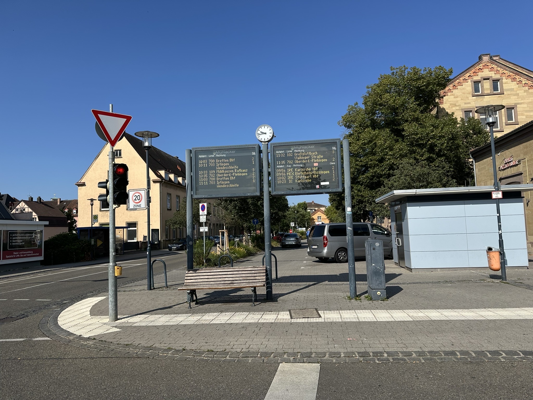 Anzeigetafel mit Abfahrtszeiten für Busse und Bahnen auf einem Platz mit einer Bank, im Hintergrund stehen Häuser und Bäume 